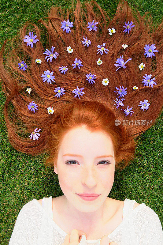 14 / 15岁的红发少女躺在草地上，皮肤苍白，脸上长着雀斑，头发散布在她的头上，点缀着白色的雏菊和紫色的花朵